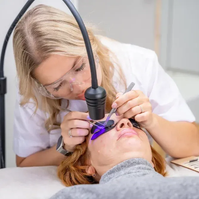 Professional beautician uses UV/LED lamp on comfortable client during eyelash extension curing treatment