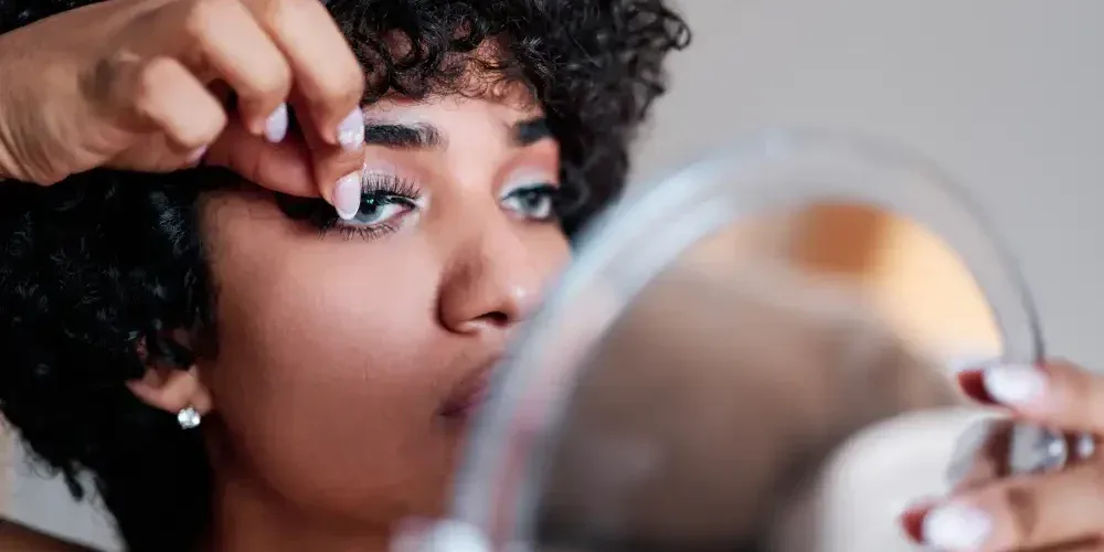 A woman attempts to apply lash segments to her own eye using a mirror, illustrating the risks and challenges of DIY lash extension kits.