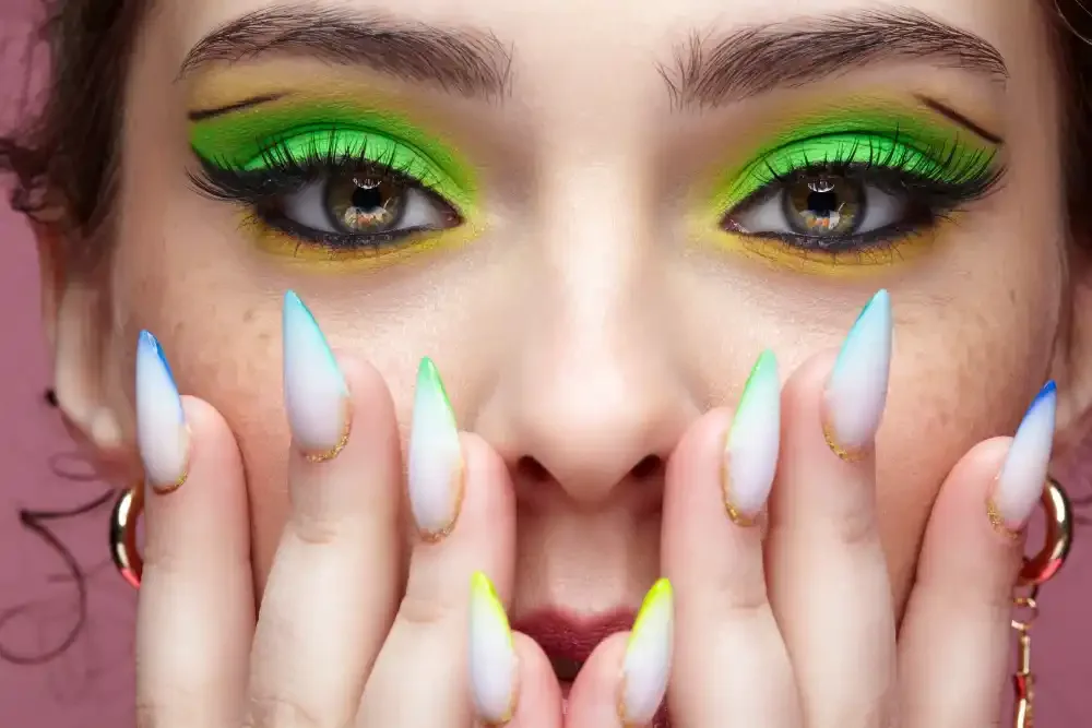 Woman with vibrant green lash extensions, bold eye makeup, and colorful pointed nails.