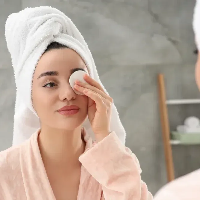 Woman with a towel around her hair gently cleansing her eye with a cotton pad.