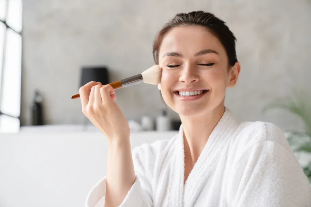 Smiling woman in a robe applying eye makeup.