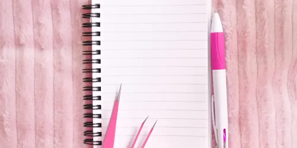 A notepad and pen next to professional lash tweezers, representing the process of creating an optimal fill schedule.