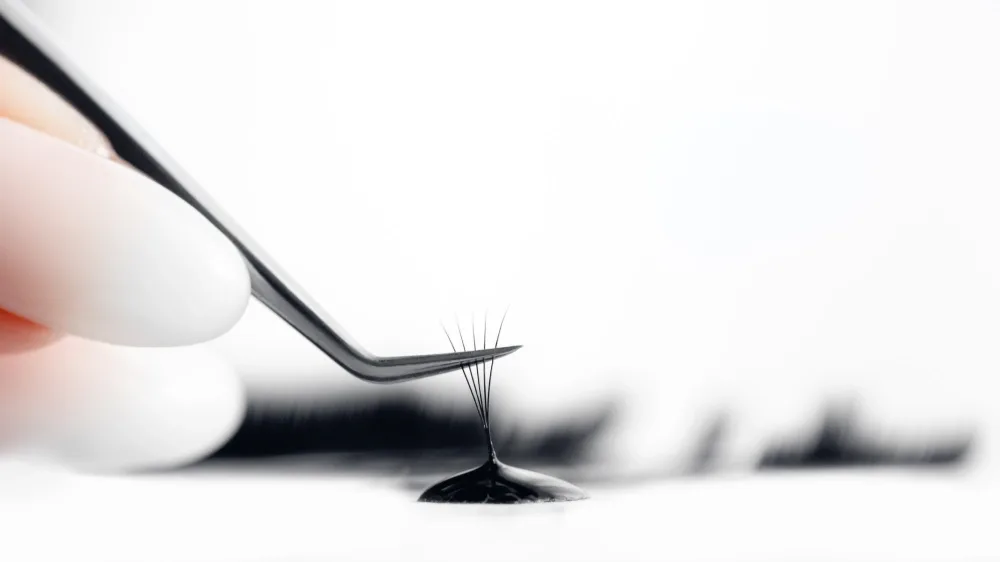 Tweezers dip a lash fan into adhesive while preparing an allergy patch test.