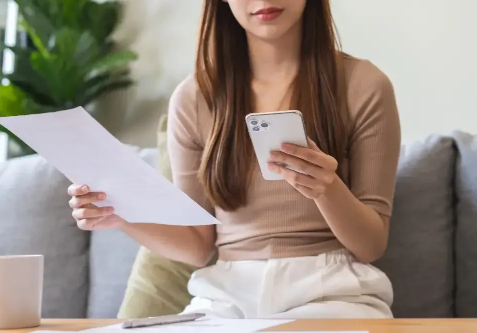 A woman compares prices on her phone and a document, illustrating the cost difference between an initial lip tattoo procedure and a later correction.
