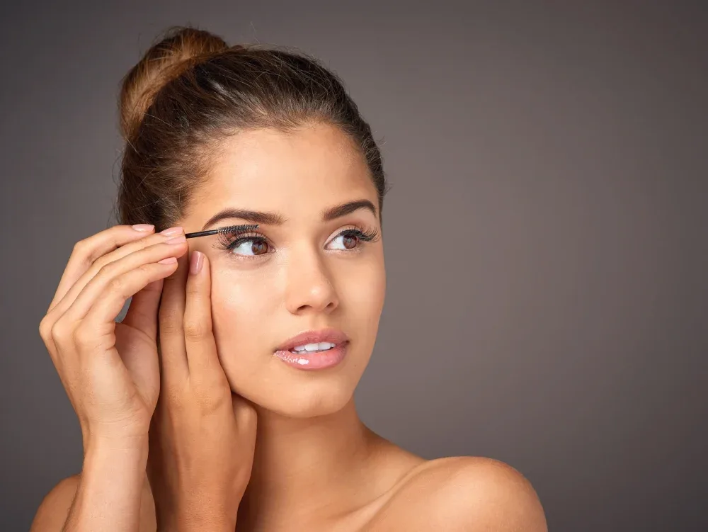 A woman carefully applies an oil-free serum to the roots of her lashes while wearing eyelash extensions to improve retention.