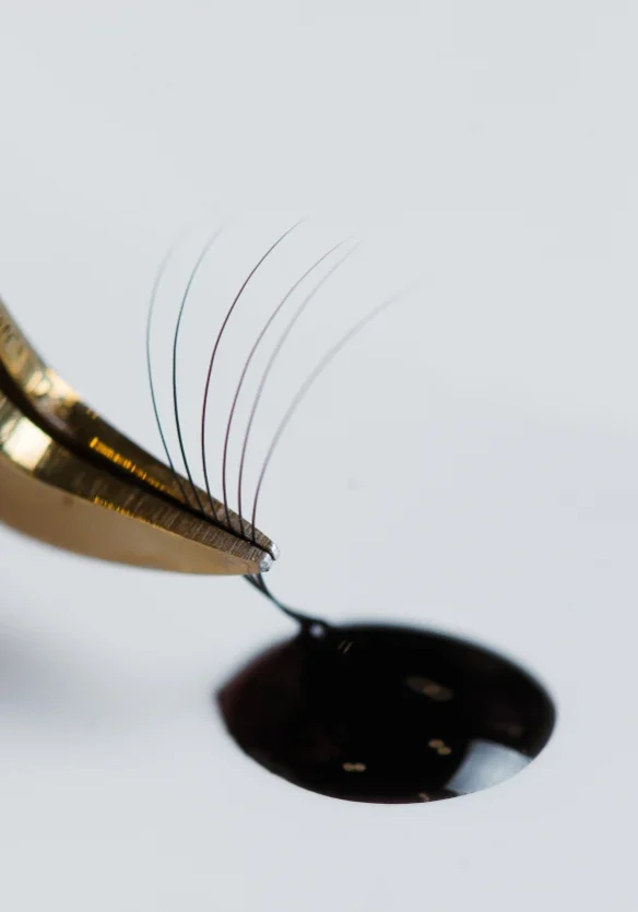 Tweezers hold a fan of lashes dipped in a drop of dark adhesive, highlighting the importance of hypoallergenic aspects of both lash materials and glue.