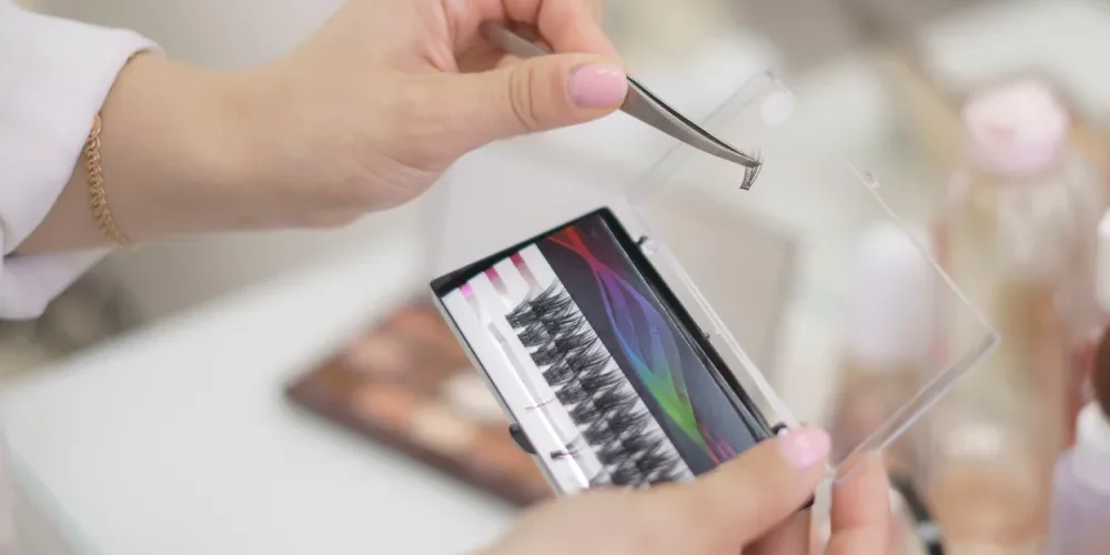 A technician selects an eyelash extension from a palette with tweezers, preparing to perform a patch test for glue allergies.