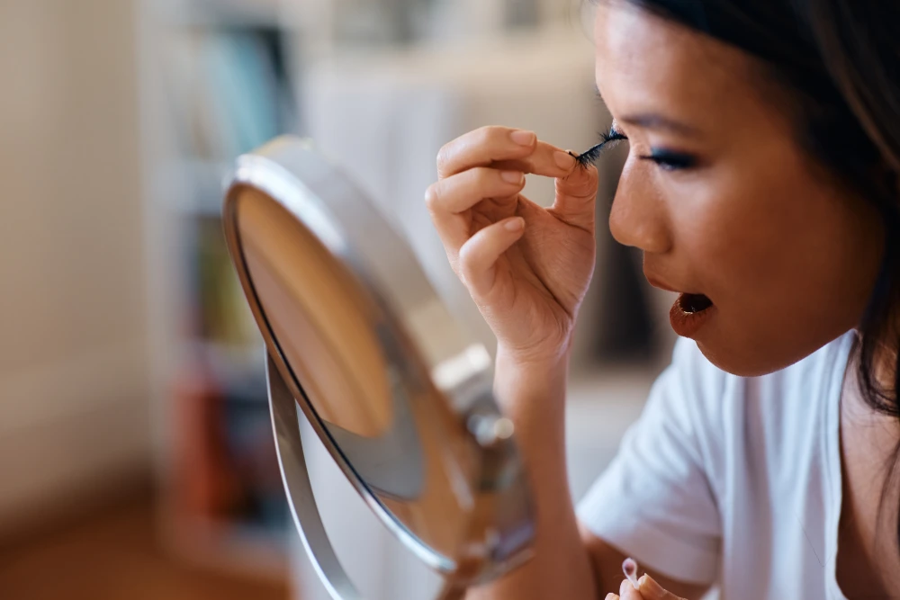 A woman struggles to remove her eyelash extensions in front of a mirror, highlighting the risks of DIY removal and potential damage.