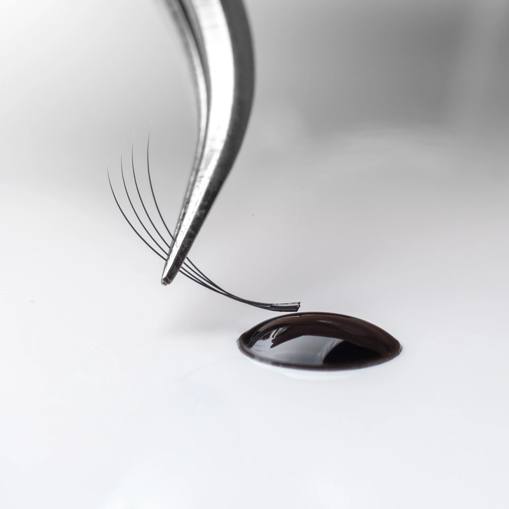 Tweezers hover over a drop of lash glue, illustrating the allergy risks associated with adhesives used in eyelash extensions, as well as potential damage and infections.