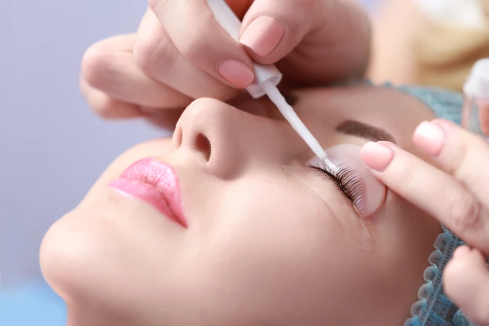 A technician applies a product to a client's eyelashes during a lamination treatment, showing the chemical effect on the natural lashes.