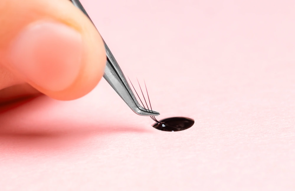 A technician uses tweezers to apply a few lash extensions, demonstrating a patch test for allergy prevention.