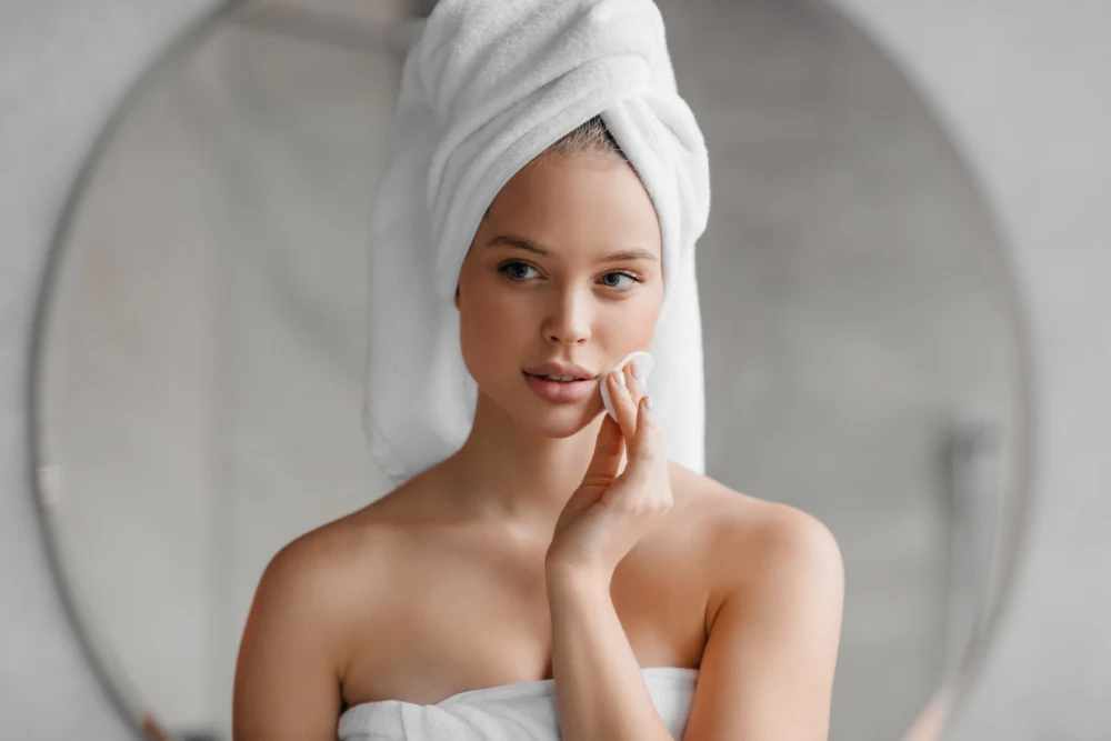 A woman with her hair in a towel cleans her face, demonstrating how to prepare for a lash extension consultation.