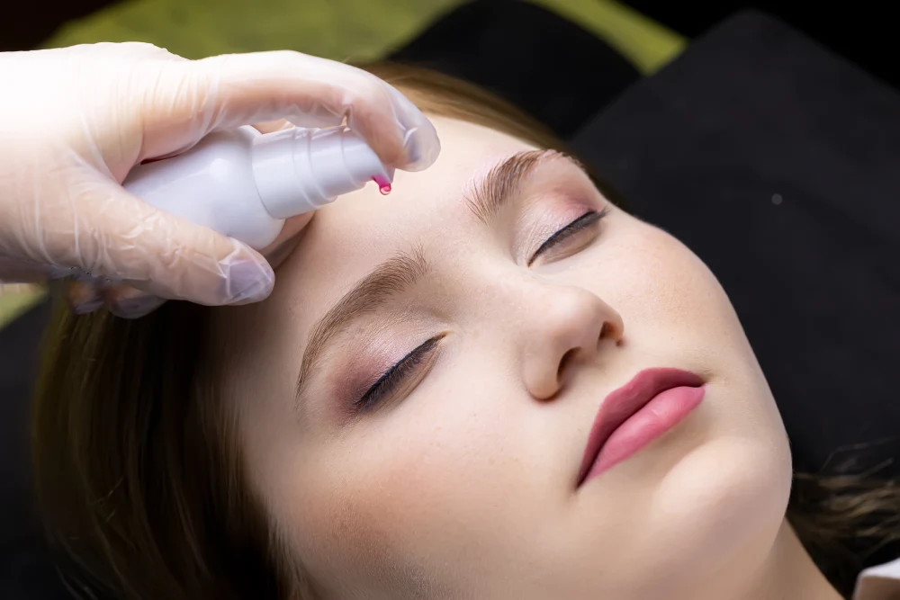A lash technician carefully cleanses a client's face with a spray bottle, preparing the eye area for an extension application.