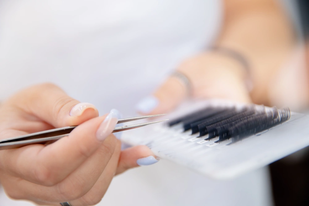 A technician uses tweezers to pick up an eyelash extension from a palette, representing the use of quality materials.