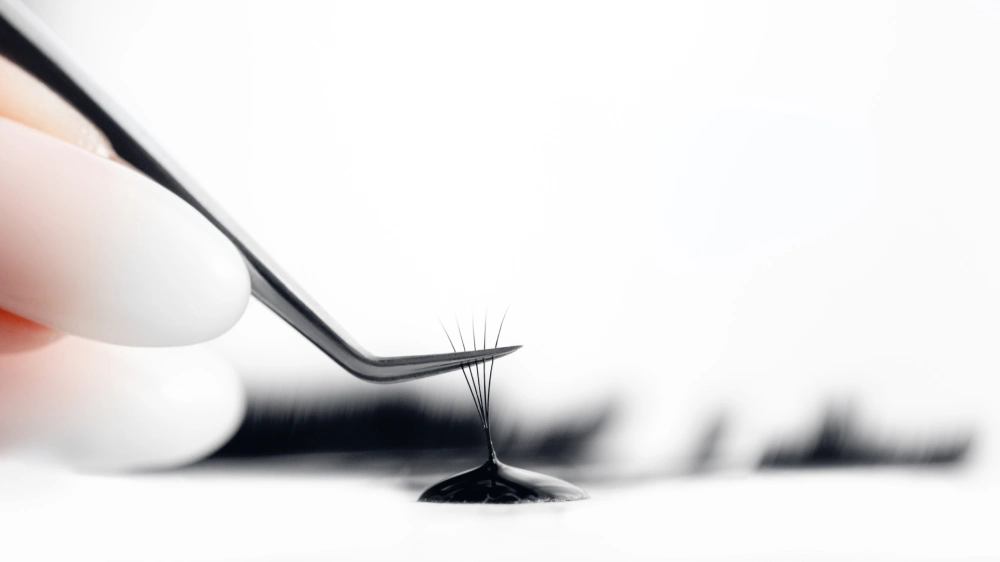 A lash fan is dipped into adhesive with tweezers, preparing for a patch test to minimize allergic reaction risks.
