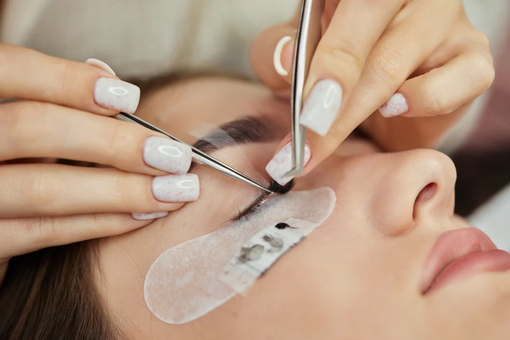 A technician gently removes softened eyelash extensions using tweezers and a micro-brush.