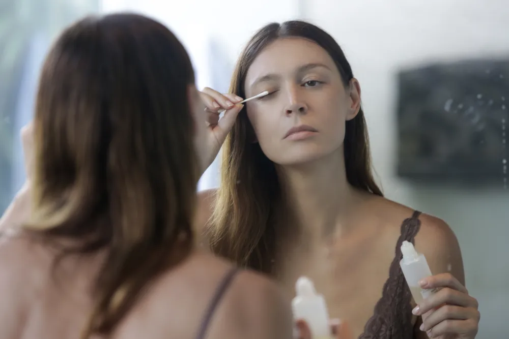 Making a Conscious Choice for Your Lashes A woman carefully applies eyelash serum while looking in a mirror, representing making a conscious choice for lash health and beauty.