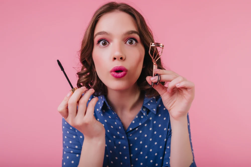 A woman with short, sparse eyelashes holds a mascara wand, illustrating the eyelash type being discussed for lamination versus extensions.